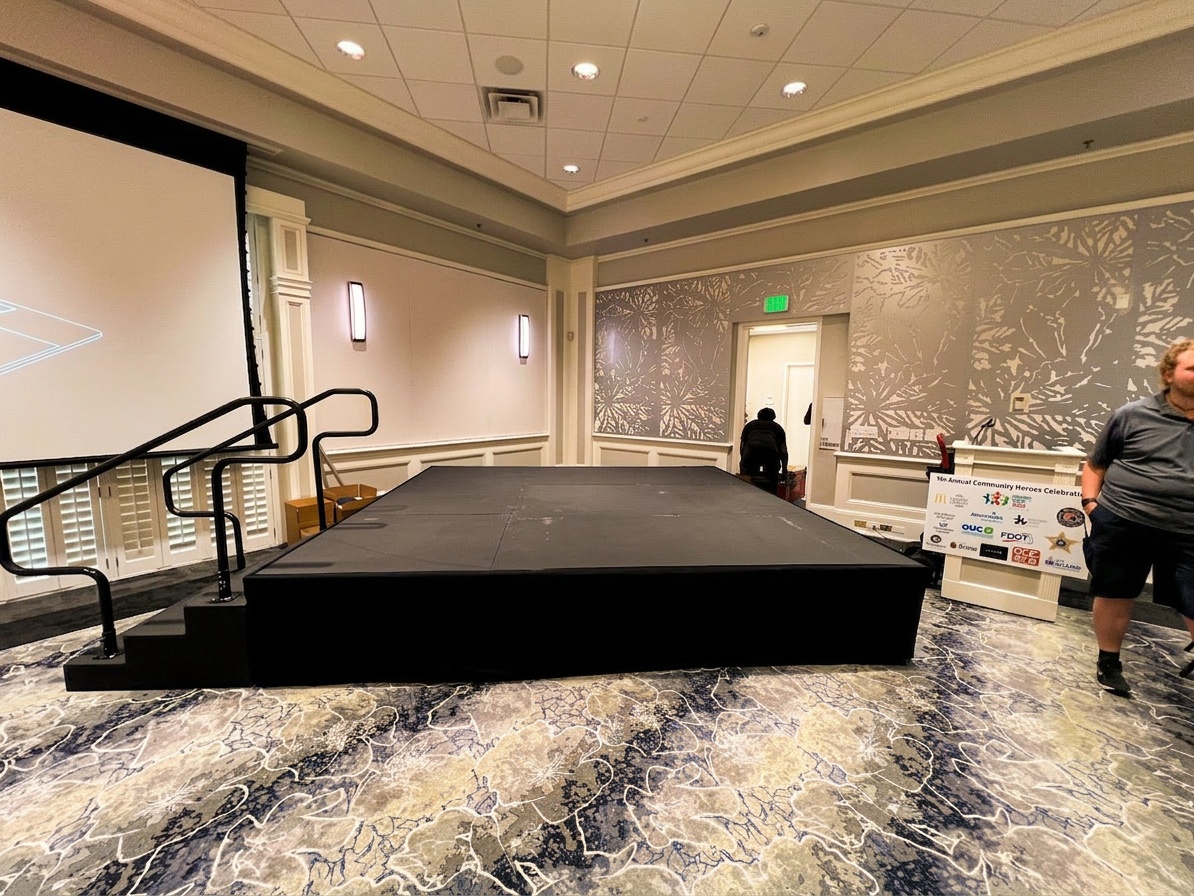 Black platform stage being set up in an elegant banquet room with decorative patterned wallpaper and a sponsor banner visible on the right side
