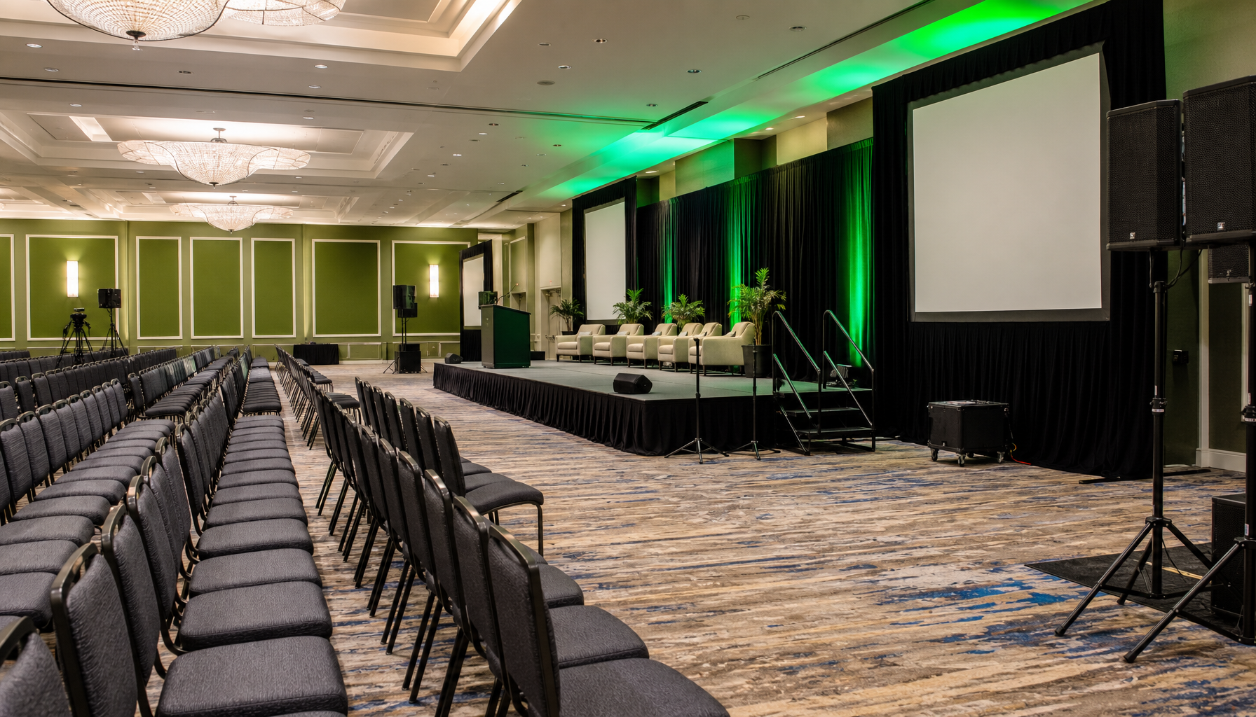 Large ballroom or conference space being set up with black stage platform and stairs, multiple chairs arranged in front, professional audio/visual equipment visible, and green accent walls in the background