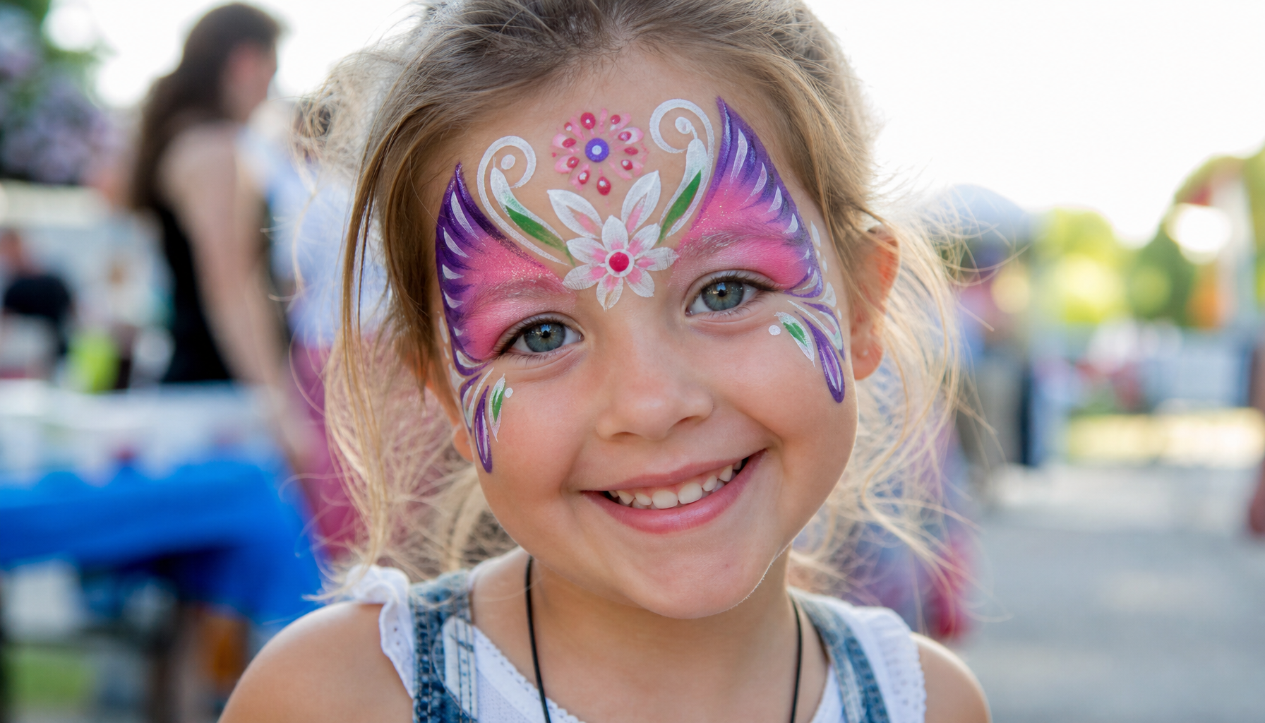 a happy child with colorful face paint at an outdoor family event, smiling face close-up showing butterfly or floral design in vibrant colors