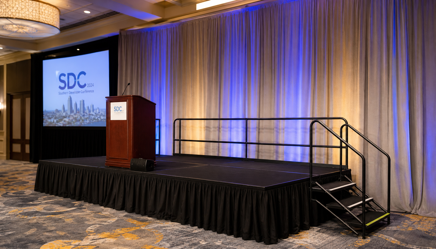 finished corporate stage with podium, black skirting, guard rails, and stair access at stage right inside a hotel ballroom